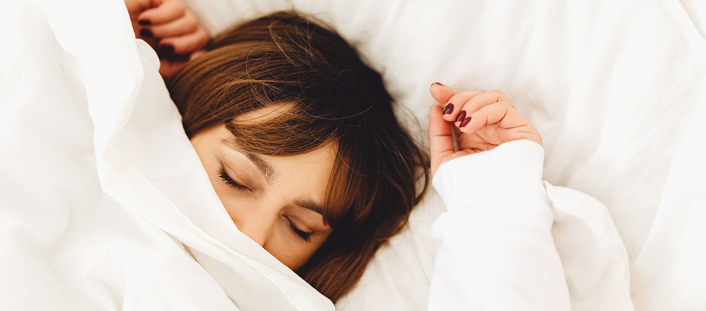 rapariga de cabelo castanho, a dormir dentro da cama de barriga para cima, com pijama comprido de cor branca, com o edredão a tapar até ao nariz