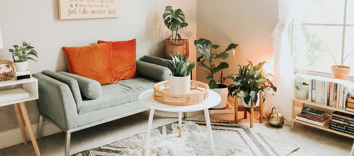 quarto com um banco de sofá com dois travesseiros, em frente uma mesinha redonda com uma bandeja com 1 vaso de flores, e ao lado 3 vasos e um móvel baixo com livros