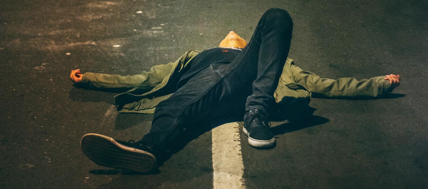 homem deitado de barriga para cima na rua, inconsciente, vestido com casaco verde, calças de ganga azuis-marinho, e sapatilhas de tecido azul escuro