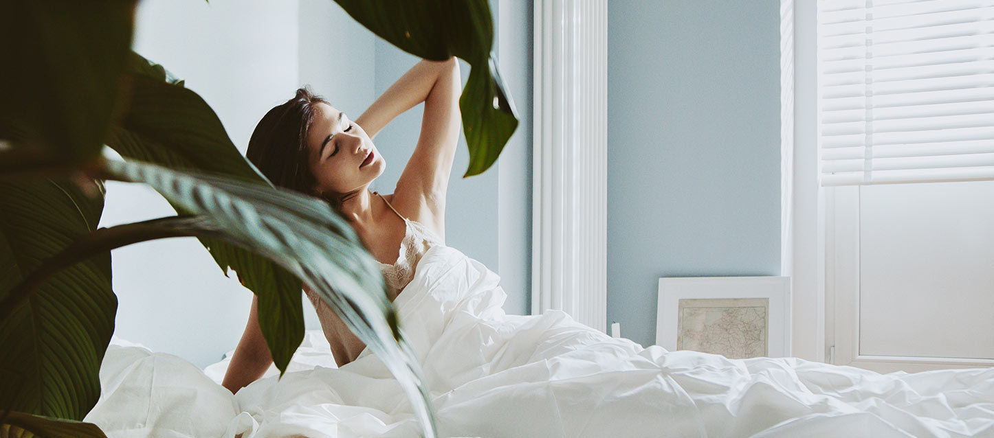rapariga em camisola a despertar-se e a meter-se na cama, coberta com edredão branco, com uma mão na nuca, e apoiando-se com o outro braço