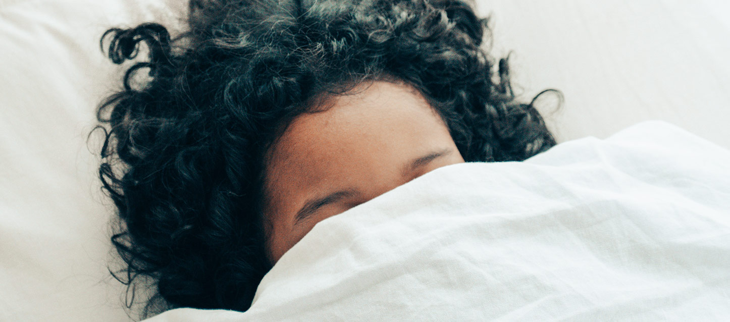 rapariga de cabelo encaracolado preto, a dormir de barriga para cima, coberta com o edredão até aos olhos