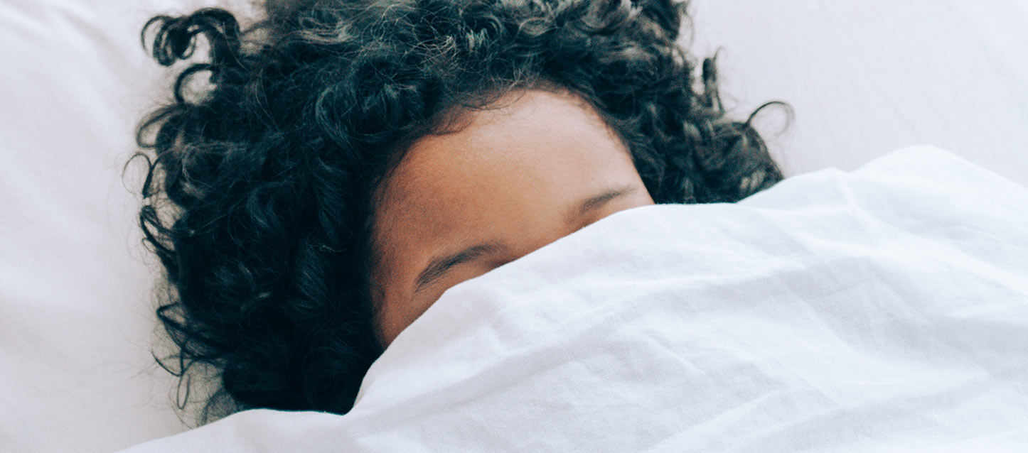 rapariga morena com o cabelo ondulado metida na cama, de barriga para cima e a dormir, e tapada com o edredão até ao nariz