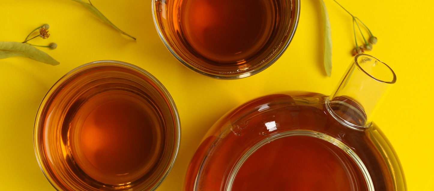 uma jarra cheia de infusão de tília, com 2 taças com infusão sobre uma mesa de cor amarela