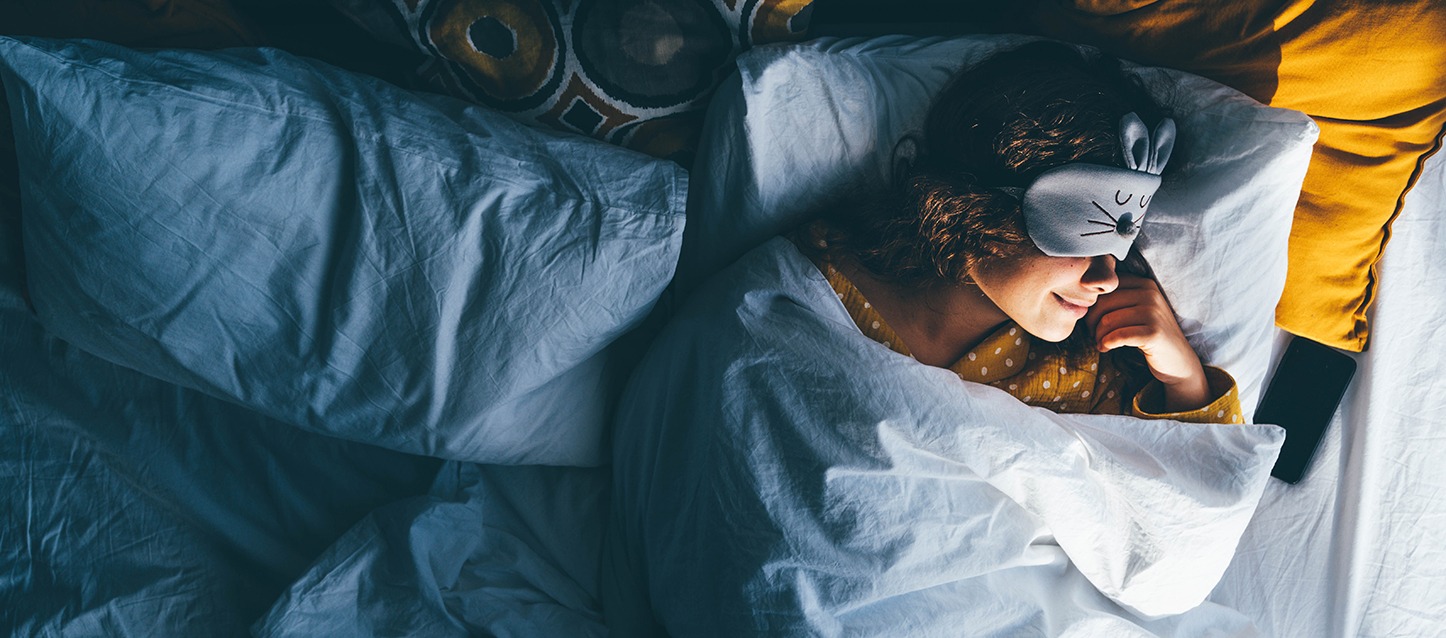 rapariga morena de cabelo encaracolado e pijama amarelo com pontos brancos, a dormir de barriga para cima e meio de lado, tapada com o edredão, e com máscara para os olhos com forma de coelho em cor cinza.