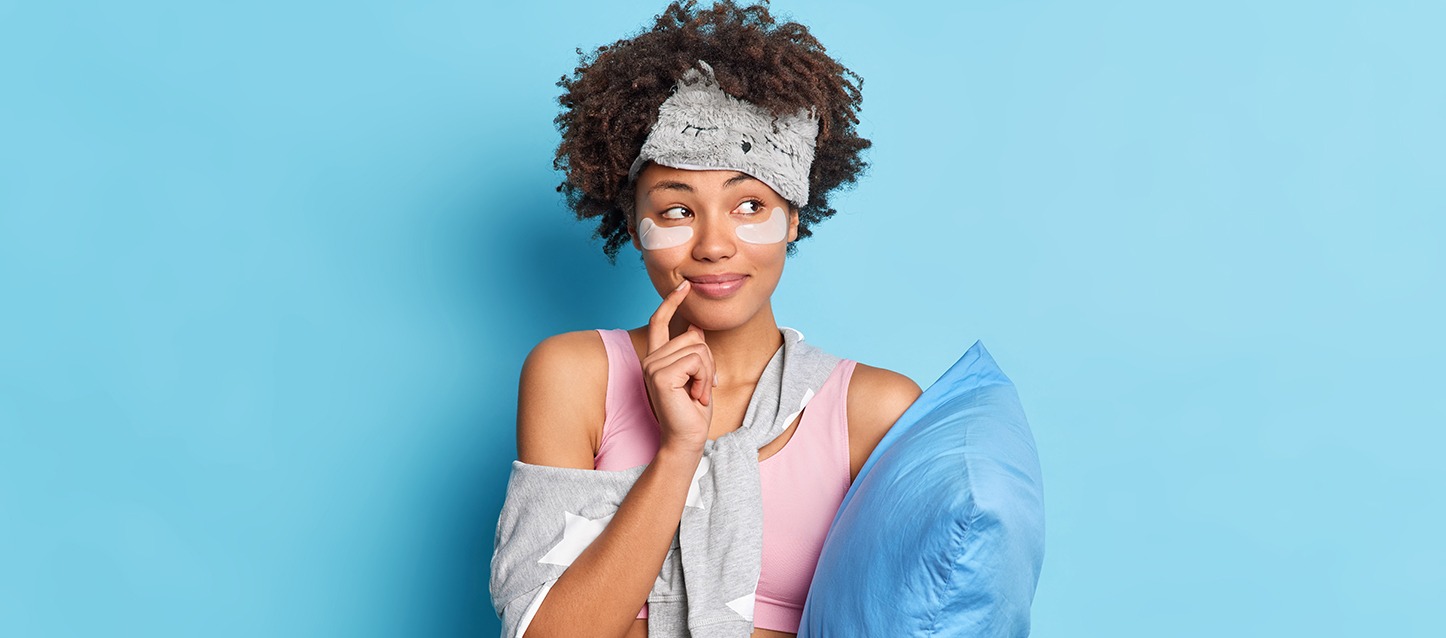rapariga morena de cabelo afro com adesivos anti-olheiras, camiseta rosa, camisola cinza com estrelas brancas aplicadas na zona do pescoço, segurando uma almofada azul, e com uma máscara para os olhos na testa