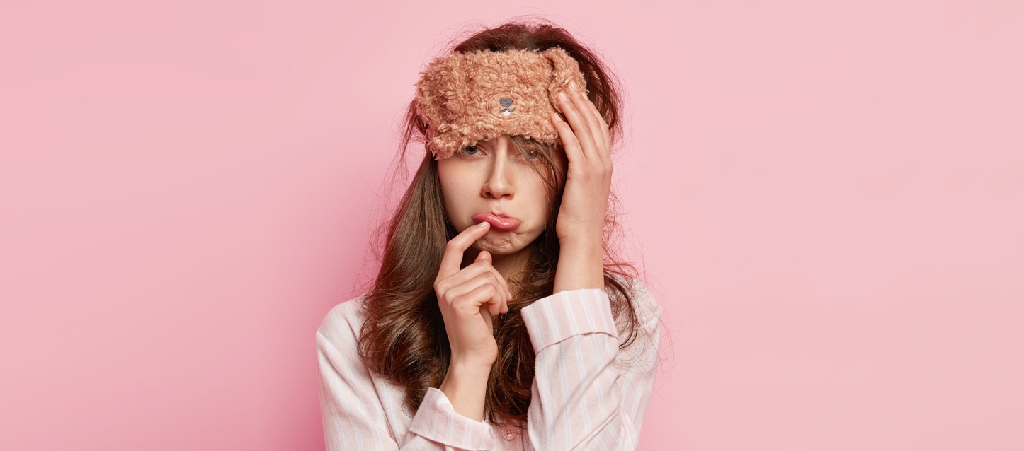 rapariga com pijama rosa e riscas brancas, máscara para os olhos com forma de cão, com uma mão na cabeça, e a outra a tocar-se o lábio com gesto cansado