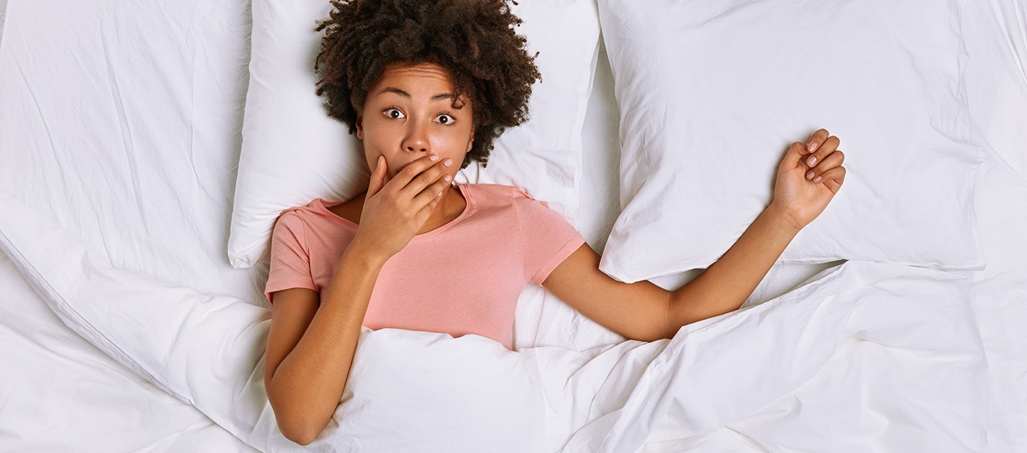 rapariga morena com cabelo afro metida na cama de barriga para cima, com camiseta rosa, e com a mão na boca em gesto de surpresa
