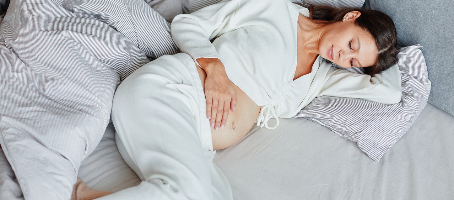 mulher grávida, de cabelo castanho escuro e pijama branco, deitada de barriga para cima e de lado, e com uma mão sobre a sua barriga