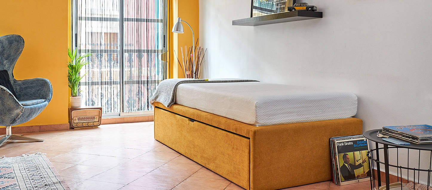cama sem cabeceira, sommier com colchão em quarto de tons amarelos, vinis apoiados no chão, e o sommier, mesinha, estante e cadeirão