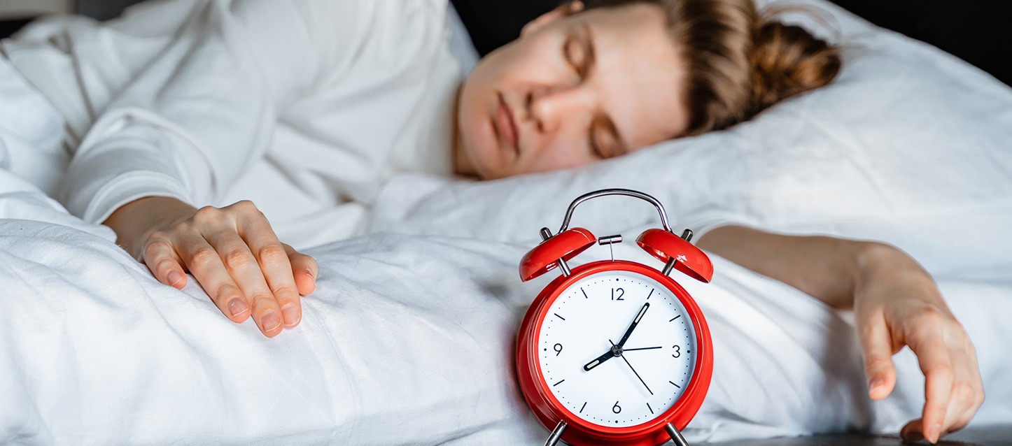 mulher dormida de lado, com pijama branco, junto a um despertador de cor vermelha