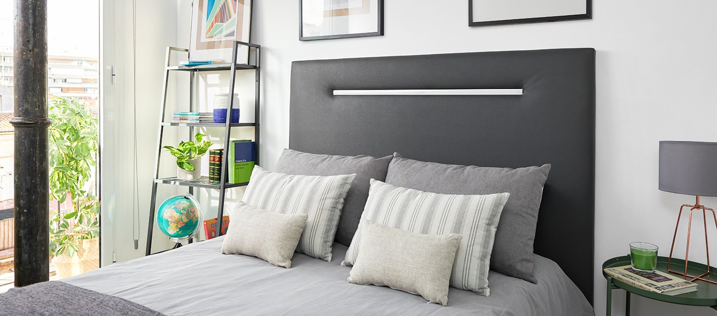 quarto pequeno decorado, cama com lençóis cinzas, 2 almofadas e 4 travesseiros, cabeceira de pele sintética, estantes com livros, mesinha e quadros