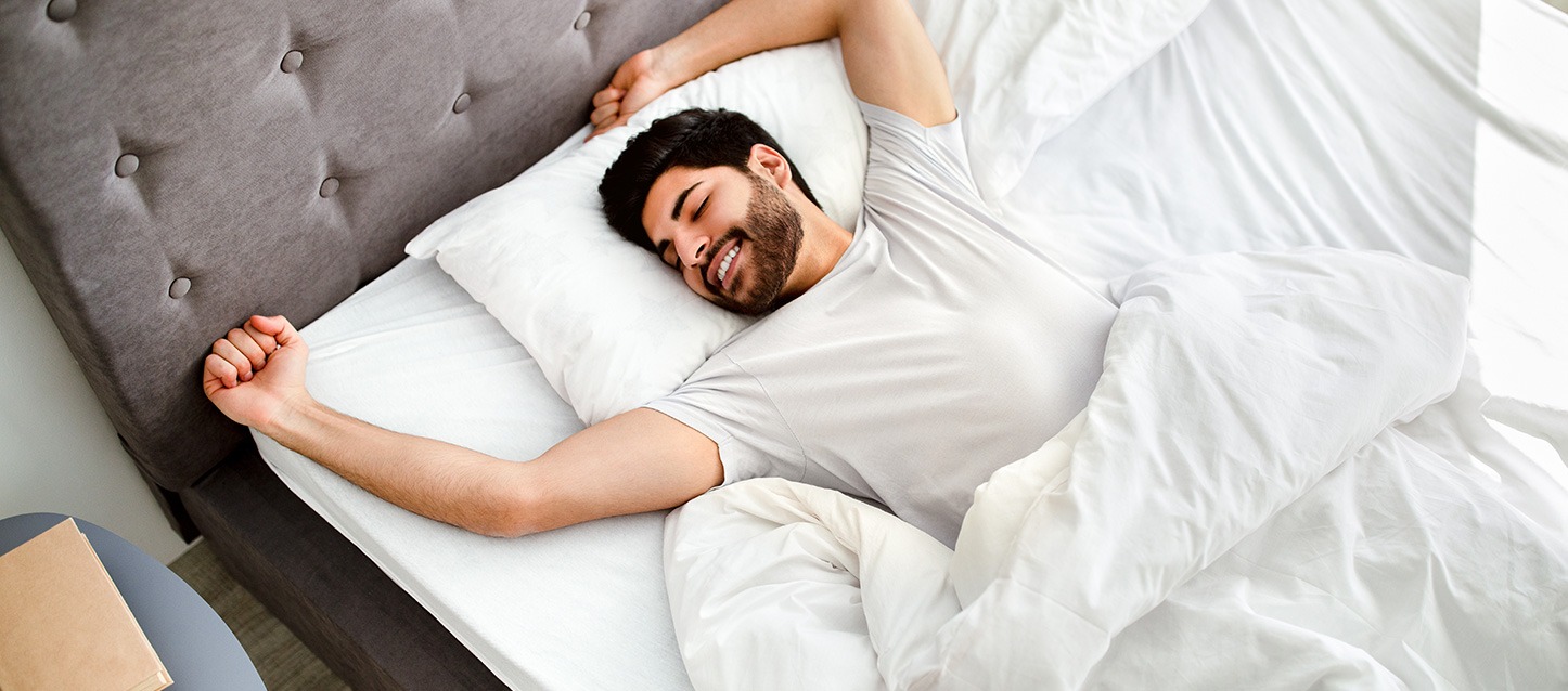 rapaz moreno com barba, deitado de barriga para cima, com camiseta branca, tapado com edredão e a espreguiçar-se
