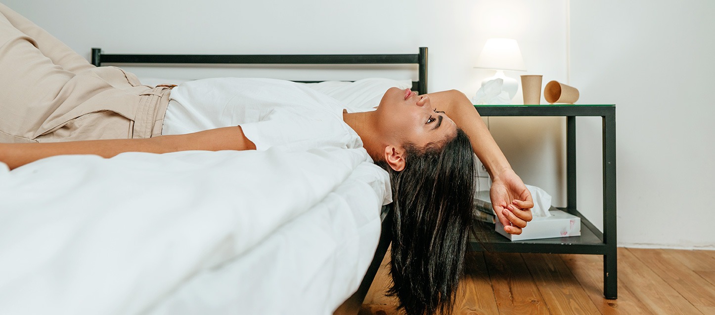 rapariga morena de cabelo comprido, camiseta branca e calça bege, deitada de barriga para cima, com a cabeça fora da cama e a olhar o teto