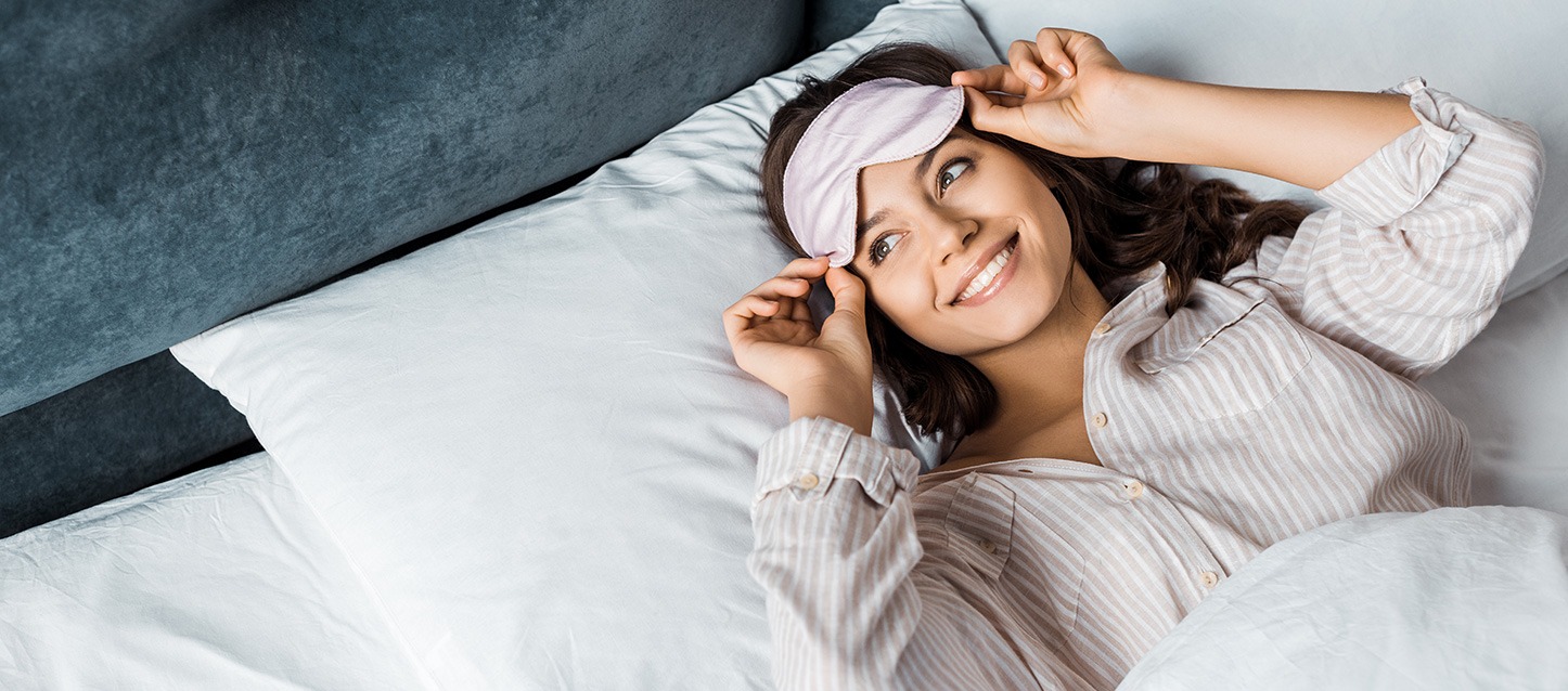 rapariga morena de cabelo comprido, metida na cama, a olhar para um lado e a sorrir enquanto remove a máscara para os olhos