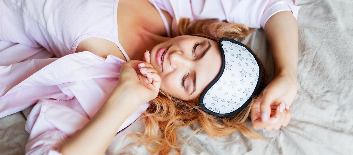 rapariga de cabelo comprido loiro, com pijama rosa e máscara para os olhos branca com estrelas cinza na testa, deitada de barriga para cima na cama