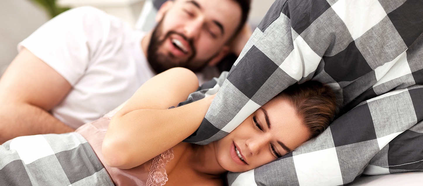 casal na cama a dormir no mesmo lado, em que ele ronca e ela se tapa a cabeça com a almofada