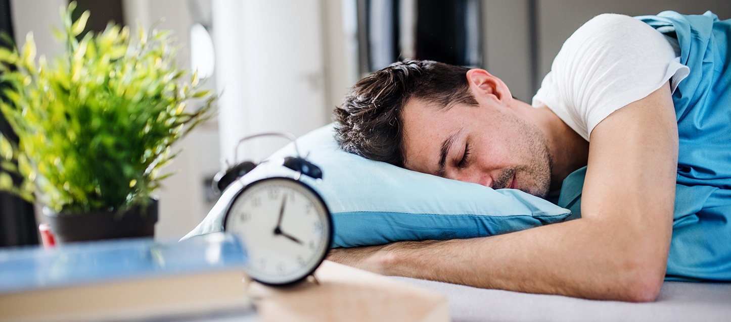 rapaz a dormir de barriga para baixo junto na cama, mesinha de cabeceira com despertador preto, e livro