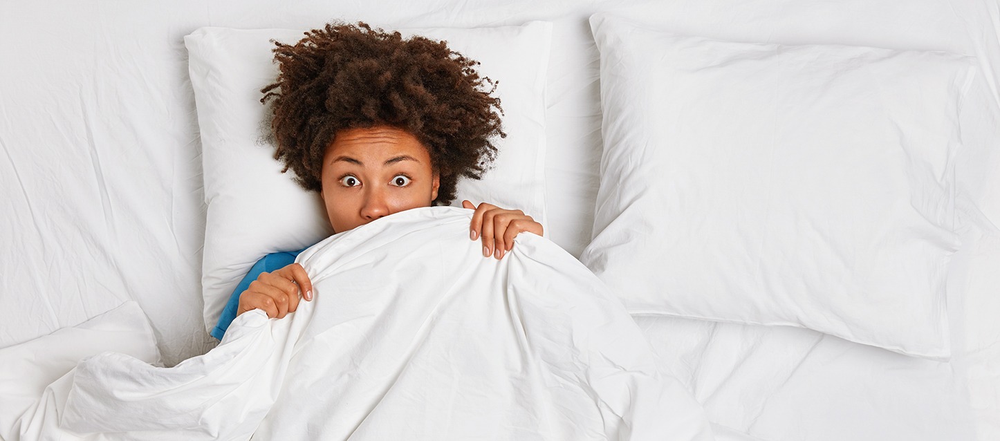 rapariga morena de cabelo afro, metida na cama, coberta com o edredão até ao nariz, com cara de assustada por ter tido pesadelos