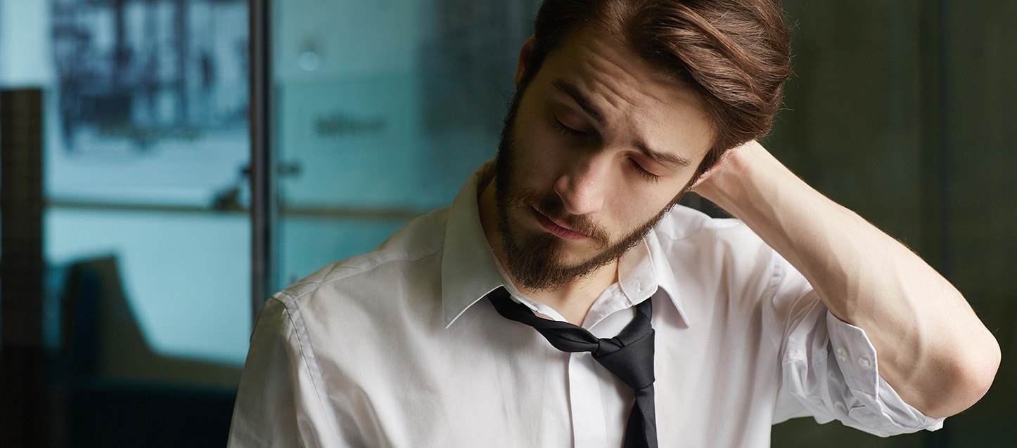 rapaz com barba, camisa branca e gravata preta, levando a mão à nuca com um gesto de dor