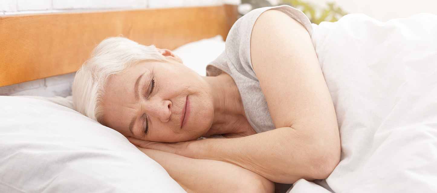 mulher idosa a dormir de lado, com as mãos juntas debaixo da cabeça, e tapada com um edredão branco
