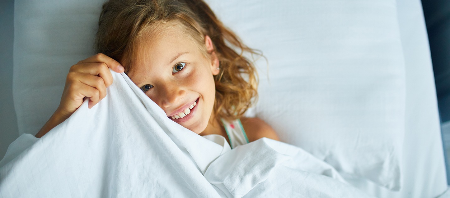 menina metida na cama, coberta com lençol branco, a puxar o lençol e a tapar um pouco a cara