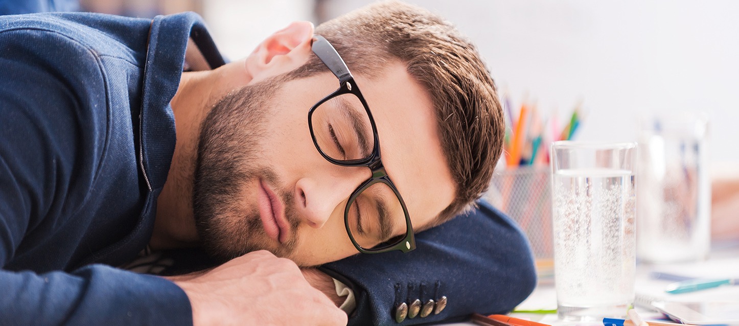 rapaz com barba, óculos e camisola azul, a dormir na mesa de trabalho em frente a um copo com água