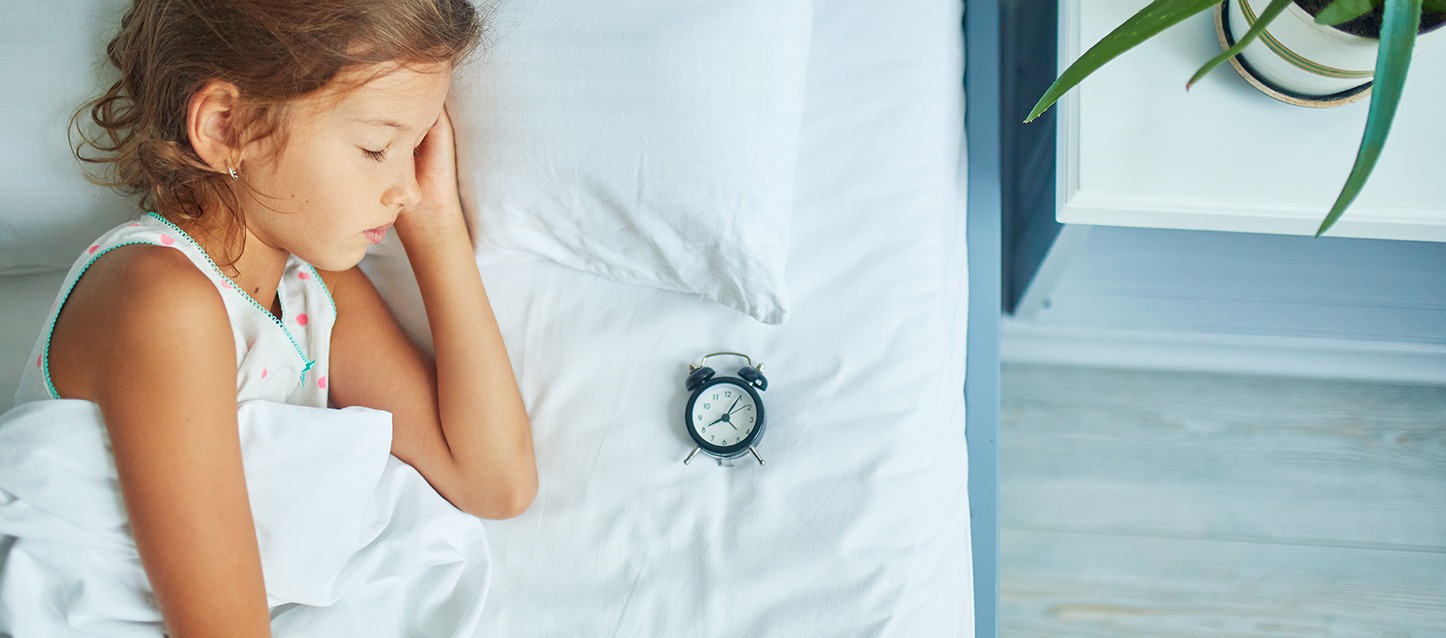 menina a dormir de lado na cama, com um despertador pequeno e preto sobre a mesma cama