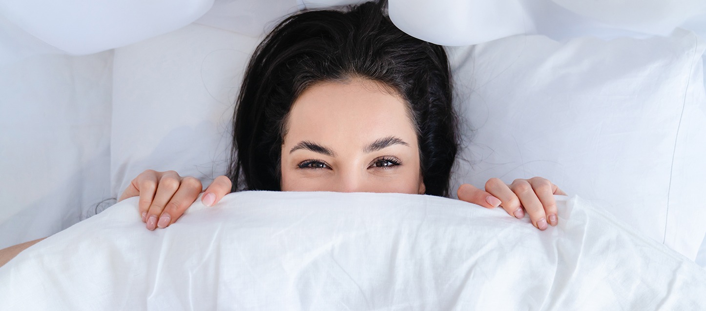 rapariga morena de cabelo comprido e olhos negros, metida na cama de barriga para cima, tapada com o edredão até ao nariz e a olhar em frente