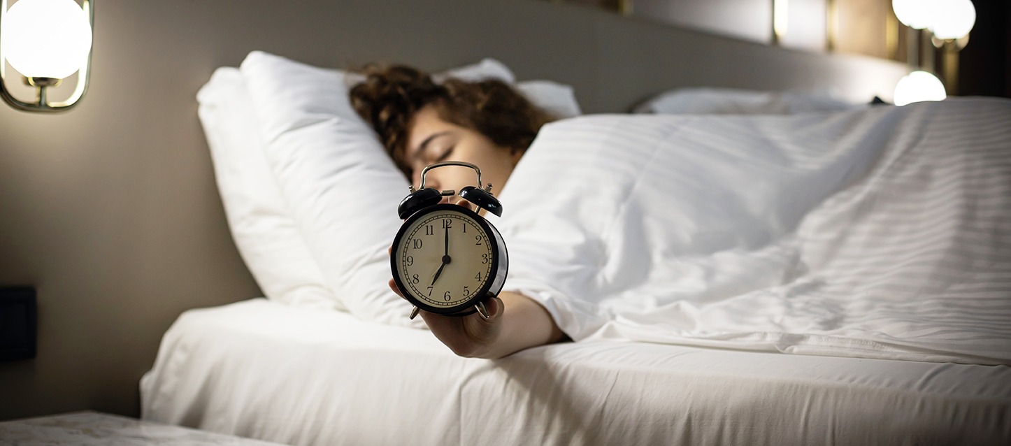 rapariga a dormir de barriga para baixo, tapada com edredão branco, e a segurar um despertador preto com a mão