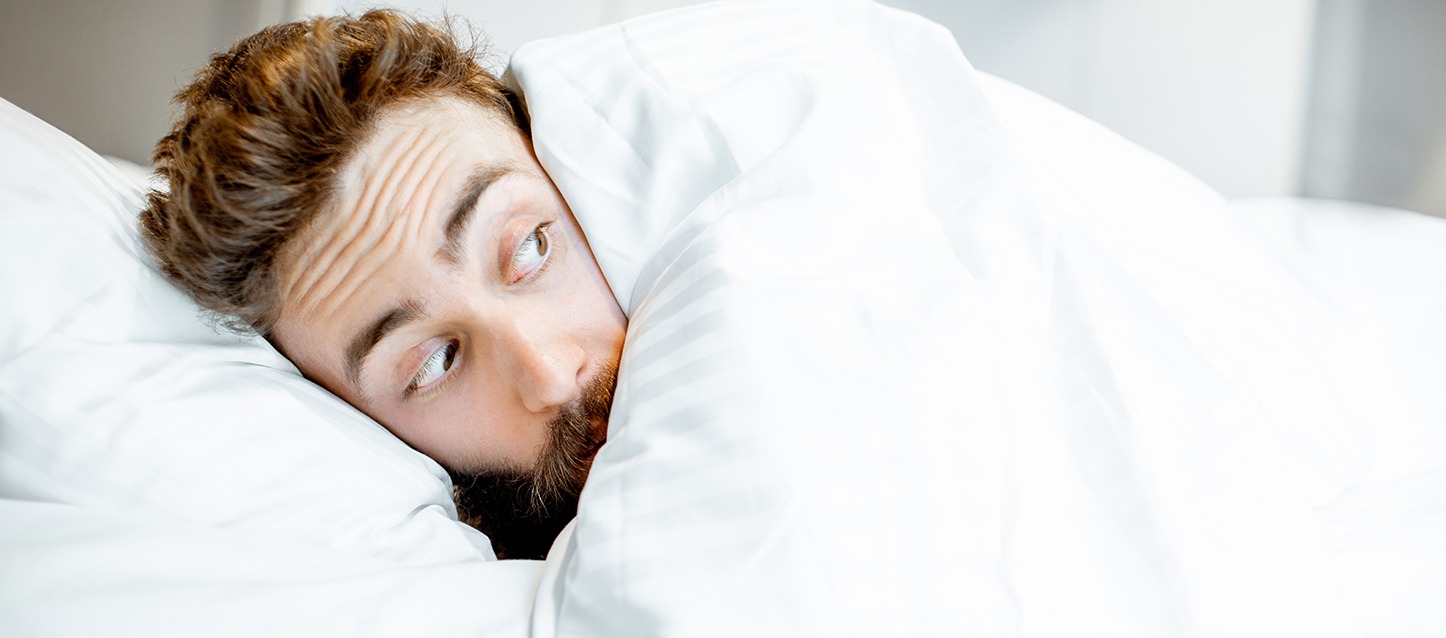 rapaz com barba metido na cama, com o edredão até à cara, a olhar com cara de susto depois de ter tido pesadelos