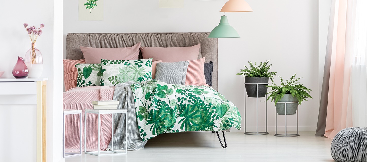 quarto com tons suaves, cabeceira castanha, vários travesseiros de tons rosa e cinza, e edredão e travesseiros brancos com motivos verdes de folhas e plantas