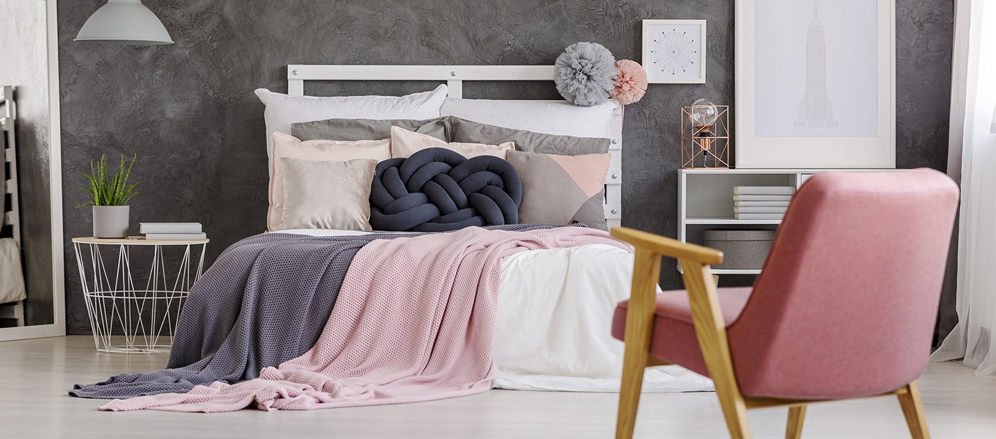 quarto com cama, 2 edredões azul e rosa, várias almofadas de diferentes tamanhos e cores, cadeira rosa, mesinha com planta e dois livros