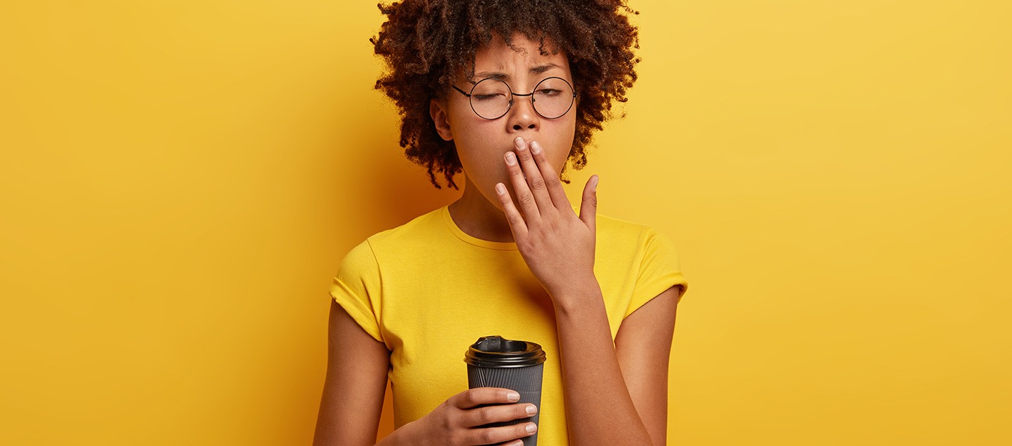 rapariga morena com óculos, de cabelo afro, a bocejar com uma mão na boca, e a segurar um copo de café com a outra