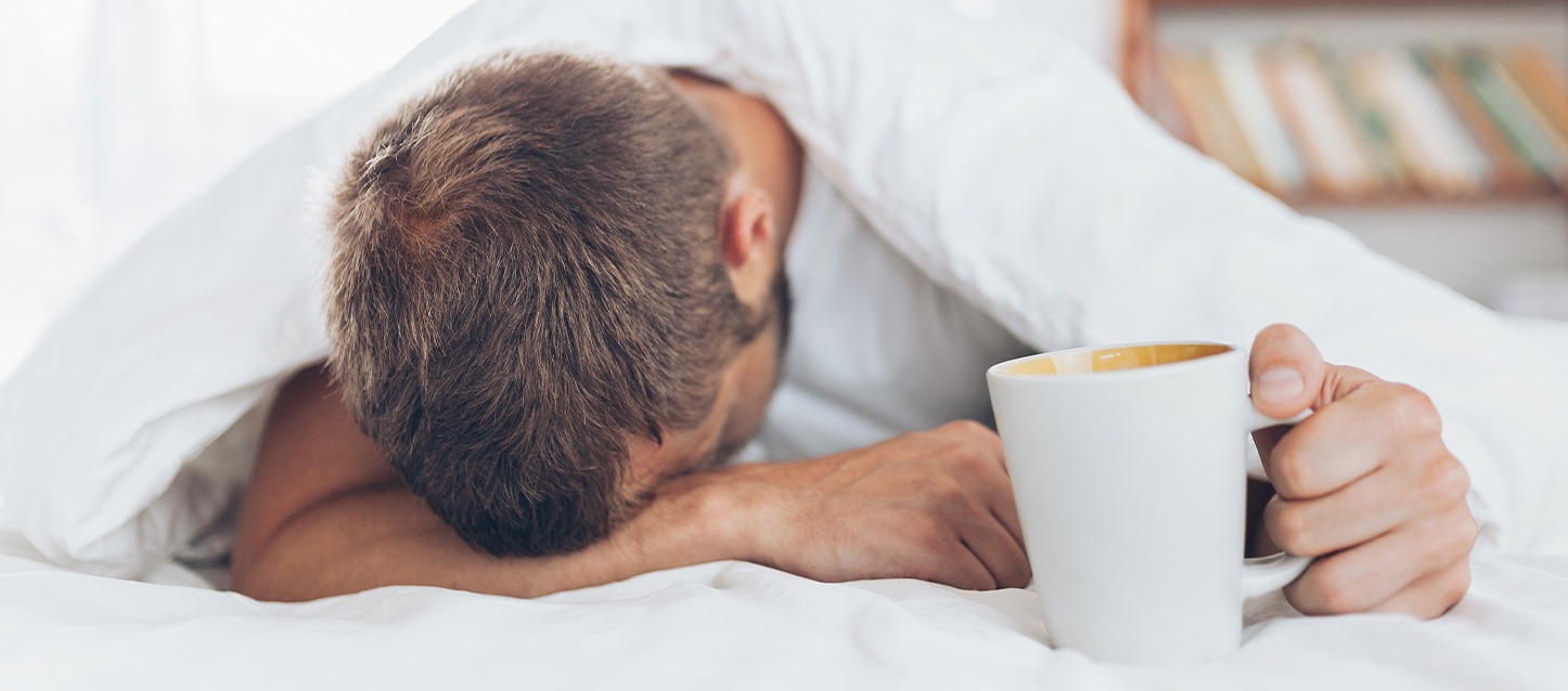 Homem deitado na cama de barriga para baixo, com a cabeça apoiada sobre o braço direito, e segurando uma taça na outra mão.