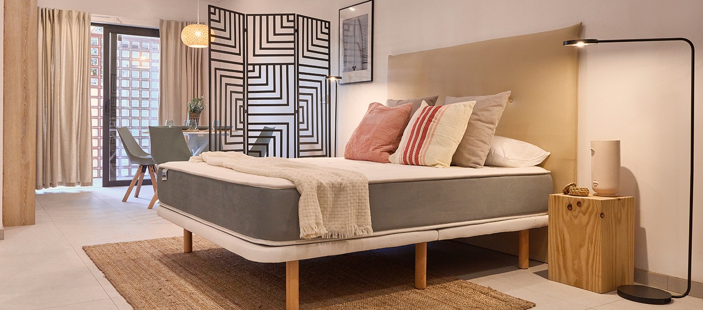 Quarto com cama, mesinha de cabeceira de madeira, mesa, cadeiras e varanda.